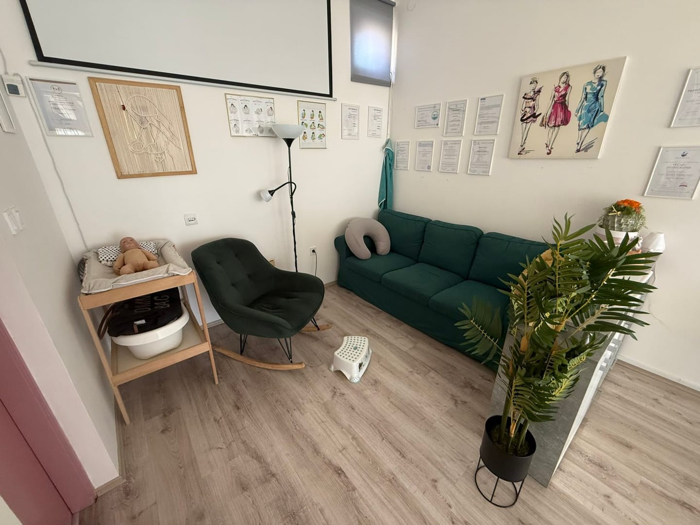 Kutak za dojenje u Prelogu: zelena sofa s jastukom za dojenje, fotelja za ljuljanje i stolić za previjanje s lutkom bebe